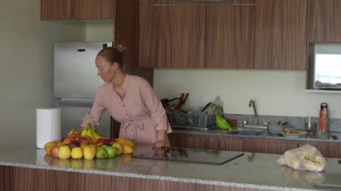 Genç bir kadın, sağlıklı ve renkli bir düzenleme hazırlamak için mutfaktaki bir masaya taze meyve koyar. Parlak ve davetkar bir yerde ev yapımı, sağIık ve düşünceli bir yaşam..