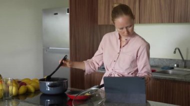 Genç bir kadın, tabletindeki videoyu izleyerek yeni bir tarif öğrenirken mutfakta yemek pişiriyor. Sıcak bir evde modern ve ilgi çekici bir mutfak keşfi, teknoloji ve yaratıcılık anı.