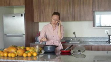 Mutfakta yemek pişirirken cep telefonuyla konuşan bir kadın. Meşgul ama rahat bir ev ortamında iletişim ve yemek hazırlama için uygun bir an.