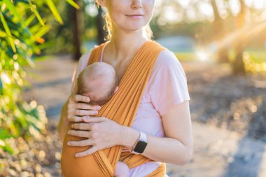 Sevgi dolu bir anne, bebeğini güvenli bir şekilde askıda taşıyarak huzurlu bir parkta yürür. Annelik bağının, bebek giyiminin ve anneliğin doğadaki neşesinin yürek ısıtan bir anı..