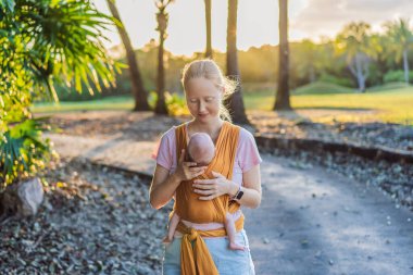 Sevgi dolu bir anne, bebeğini güvenli bir şekilde askıda taşıyarak huzurlu bir parkta yürür. Annelik bağının, bebek giyiminin ve anneliğin doğadaki neşesinin yürek ısıtan bir anı..
