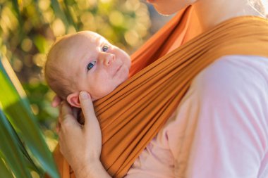Sevgi dolu bir anne, bebeğini güvenli bir şekilde askıda taşıyarak huzurlu bir parkta yürür. Annelik bağının, bebek giyiminin ve anneliğin doğadaki neşesinin yürek ısıtan bir anı..