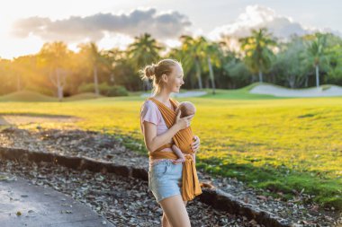 Sevgi dolu bir anne, bebeğini güvenli bir şekilde askıda taşıyarak huzurlu bir parkta yürür. Annelik bağının, bebek giyiminin ve anneliğin doğadaki neşesinin yürek ısıtan bir anı..