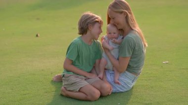 Genç bir anne, yemyeşil bir çimenlikte büyük oğlu ve yeni doğmuş çocuğuyla güneşli bir günün tadını çıkarır. Sevgi, bağ ve açık hava eğlencesiyle dolu neşeli bir aile anı ebeveynliği ve çocukluğu kutluyor.