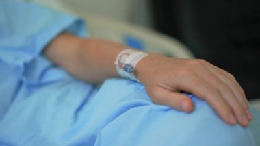 Hastanedeki bir tıp yatağında dinlenirken damar içi sondayla bir kadının elinin yakın çekimi. Klinik ortamda tıbbi bakım, iyileşme ve hasta tedavisini yansıtan detaylı bir görünüm
