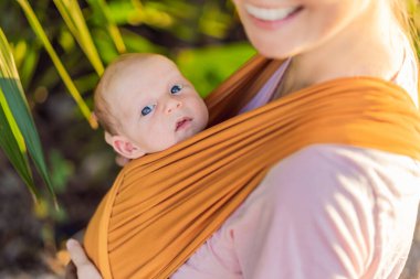 Sevgi dolu bir anne, bebeğini güvenli bir şekilde askıda taşıyarak huzurlu bir parkta yürür. Annelik bağının, bebek giyiminin ve anneliğin doğadaki neşesinin yürek ısıtan bir anı..