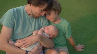 Genç bir baba, yemyeşil bir çimenlikte büyük oğlu ve yeni doğmuş çocuğuyla güneşli bir günün tadını çıkarır. Sevgi, bağ ve açık hava eğlencesiyle dolu neşeli bir aile anı ebeveynliği ve çocukluğu kutluyor.
