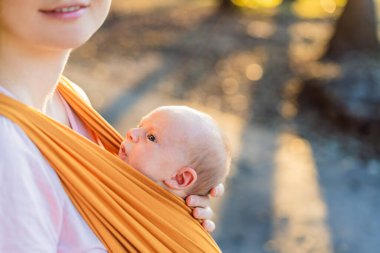Sevgi dolu bir anne, bebeğini güvenli bir şekilde askıda taşıyarak huzurlu bir parkta yürür. Annelik bağının, bebek giyiminin ve anneliğin doğadaki neşesinin yürek ısıtan bir anı..