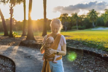 Sevgi dolu bir anne, bebeğini güvenli bir şekilde askıda taşıyarak huzurlu bir parkta yürür. Annelik bağının, bebek giyiminin ve anneliğin doğadaki neşesinin yürek ısıtan bir anı..