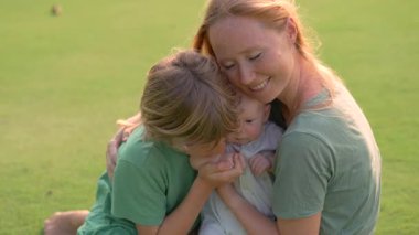 Genç bir anne, yemyeşil bir çimenlikte büyük oğlu ve yeni doğmuş çocuğuyla güneşli bir günün tadını çıkarır. Sevgi, bağ ve açık hava eğlencesiyle dolu neşeli bir aile anı ebeveynliği ve çocukluğu kutluyor.