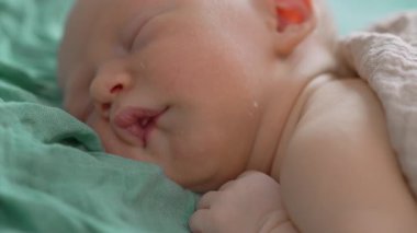 Uyuyan yeni doğmuş bir bebeğin yumuşak bir muslin battaniyeye sarılı olduğu yakın çekim videosu. Yumuşak ve huzurlu bir an, yeni doğmuş bir bebeğin masumiyetini ve huzurunu yakalıyor.