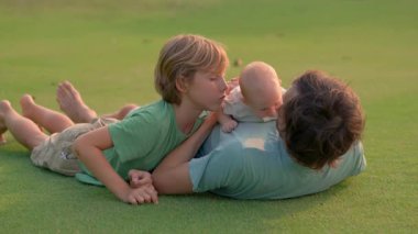 Genç bir baba, yemyeşil bir çimenlikte büyük oğlu ve yeni doğmuş çocuğuyla güneşli bir günün tadını çıkarır. Sevgi, bağ ve açık hava eğlencesiyle dolu neşeli bir aile anı ebeveynliği ve çocukluğu kutluyor.