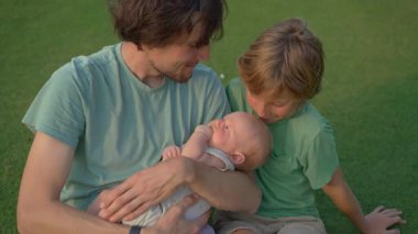 Genç bir baba, yemyeşil bir çimenlikte büyük oğlu ve yeni doğmuş çocuğuyla güneşli bir günün tadını çıkarır. Sevgi, bağ ve açık hava eğlencesiyle dolu neşeli bir aile anı ebeveynliği ve çocukluğu kutluyor.