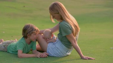 Genç bir anne, yemyeşil bir çimenlikte büyük oğlu ve yeni doğmuş çocuğuyla güneşli bir günün tadını çıkarır. Sevgi, bağ ve açık hava eğlencesiyle dolu neşeli bir aile anı ebeveynliği ve çocukluğu kutluyor.