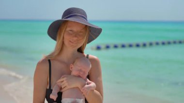 Anne ve yeni doğan oğlu güneşli bir yaz gününde kumsalda. Sevgi dolu bir annenin bebeğini sıcak gün ışığında kucağına aldığı yakın plan. Ebeveynlik, yaz havası ve hassas aile anları konsepti..
