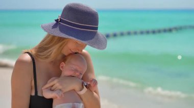 Anne ve yeni doğan oğlu güneşli bir yaz gününde kumsalda. Sevgi dolu bir annenin bebeğini sıcak gün ışığında kucağına aldığı yakın plan. Ebeveynlik, yaz havası ve hassas aile anları konsepti..