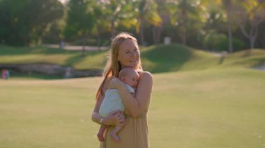 Genç bir kadın yemyeşil bir çimenlikte duruyor, yeni doğmuş bebeğini ellerinde tutuyor. Doğayla çevrili neşeli ve hassas bir bağ, aşk ve mutluluk anı.