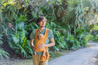 Babam küçük bebeğini parkta sarı bir askıda tutuyor. Sıcak ve sevgi dolu bir aile anı. Bebek giyimi, babalık ve ebeveyn-çocuk bağlama kavramı..