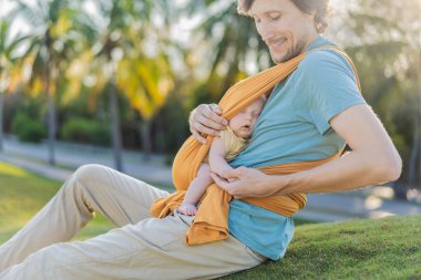 Babam küçük bebeğini parkta sarı bir askıda tutuyor. Sıcak ve sevgi dolu bir aile anı. Bebek giyimi, babalık ve ebeveyn-çocuk bağlama kavramı..