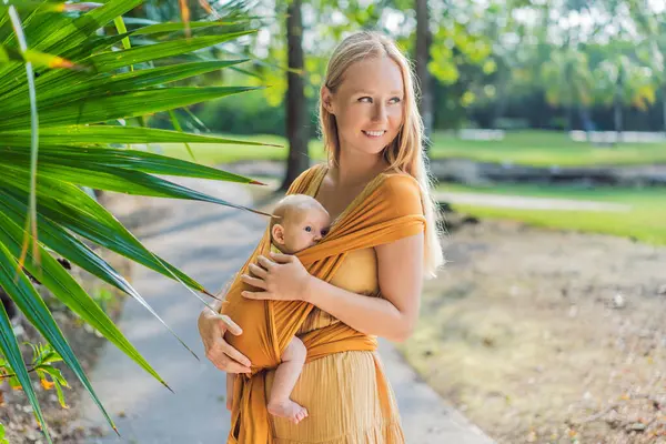 Anne, küçük bebeğini parkta sarı bir askıda tutuyor. Sıcak ve sevgi dolu bir aile anı. Bebek giyimi, ebeveynlik ve anne-çocuk bağlama kavramı.
