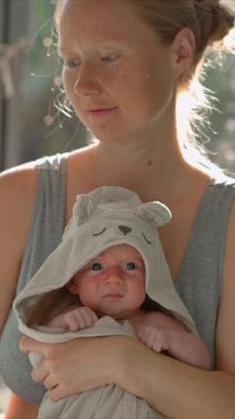 Genç bir anne sıcak bir yaz gününde güneşli bir pencerenin önünde dururken yeni doğmuş bebeğini nazikçe havluya sarar. Huzurlu ve hassas bir an. Sevgi, özen ve doğal ışıkta kaynaşma..