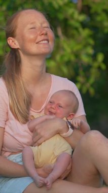 Genç bir kadın yemyeşil bir çimenlikte gülerek oturuyor, yeni doğmuş bebeğini elinde tutuyor. Doğayla çevrili neşeli ve hassas bir bağ, aşk ve mutluluk anı.