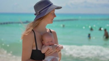 Anne ve yeni doğan oğlu güneşli bir yaz gününde kumsalda. Sevgi dolu bir annenin bebeğini sıcak gün ışığında kucağına aldığı yakın plan. Ebeveynlik, yaz havası ve hassas aile anları konsepti..