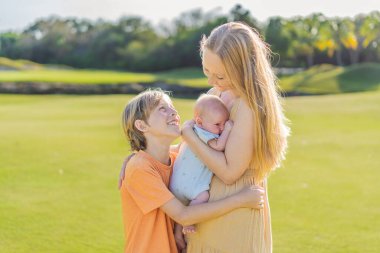 Anne, büyük oğul ve parkta bir çayırda yeni doğmuş bebek. Aile, doğada birlikte kaliteli zaman geçirir. Ebeveynlik, sevgi ve aile bağı kavramı.