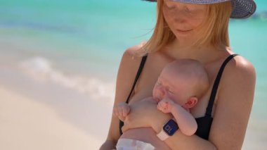 Anne ve yeni doğan oğlu güneşli bir yaz gününde kumsalda. Sevgi dolu bir annenin bebeğini sıcak gün ışığında kucağına aldığı yakın plan. Ebeveynlik, yaz havası ve hassas aile anları konsepti..