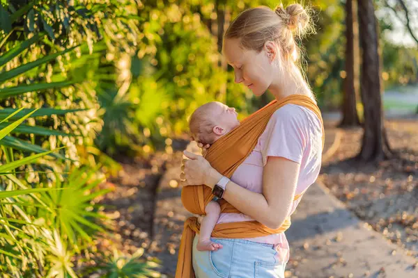 Sevgi dolu bir anne, bebeğini güvenli bir şekilde askıda taşıyarak huzurlu bir parkta yürür. Annelik bağının, bebek giyiminin ve anneliğin doğadaki neşesinin yürek ısıtan bir anı..