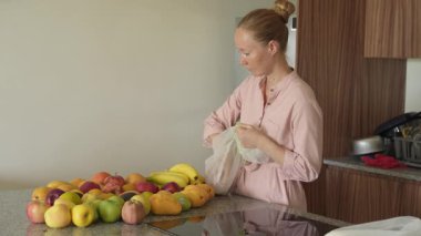 Genç bir kadın, sağlıklı ve renkli bir düzenleme hazırlamak için mutfaktaki bir masaya taze meyve koyar. Parlak ve davetkar bir yerde ev yapımı, sağIık ve düşünceli bir yaşam..