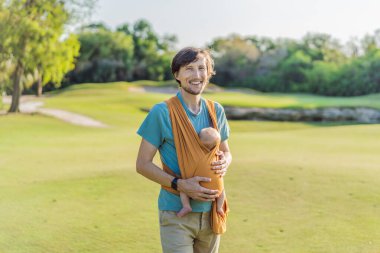 Babam küçük bebeğini parkta sarı bir askıda tutuyor. Sıcak ve sevgi dolu bir aile anı. Bebek giyimi, babalık ve ebeveyn-çocuk bağlama kavramı..
