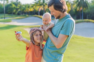 Baba ve iki oğlu parkta, büyük çocuk pilot şapkası takıyor ve bebek izlerken oyuncak bir uçak tutuyor. Çocukluk, bağ ve aile macerası konsepti.