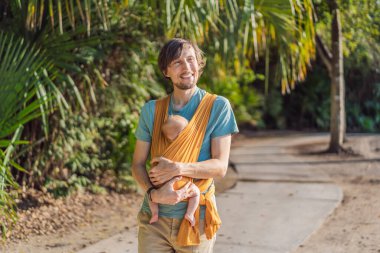 Babam küçük bebeğini parkta sarı bir askıda tutuyor. Sıcak ve sevgi dolu bir aile anı. Bebek giyimi, babalık ve ebeveyn-çocuk bağlama kavramı..