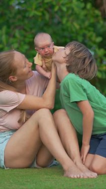 Genç bir kadın yemyeşil bir çimenlikte, yanında büyük oğlu, yeni doğmuş bebeğini ellerinde tutarken gülümsüyor. Huzurlu bir ortamda aile bağının, sevginin ve sevincin yürek ısıtan anı.