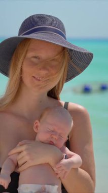 Anne ve yeni doğan oğlu güneşli bir yaz gününde kumsalda. Sevgi dolu bir annenin bebeğini sıcak gün ışığında kucağına aldığı yakın plan. Ebeveynlik, yaz havası ve hassas aile anları konsepti..