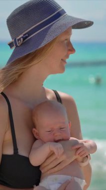 Anne ve yeni doğan oğlu güneşli bir yaz gününde kumsalda. Sevgi dolu bir annenin bebeğini sıcak gün ışığında kucağına aldığı yakın plan. Ebeveynlik, yaz havası ve hassas aile anları konsepti..