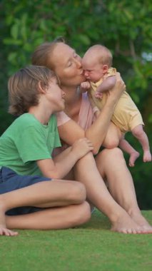 Genç bir kadın yemyeşil bir çimenlikte, yanında büyük oğlu, yeni doğmuş bebeğini ellerinde tutarken gülümsüyor. Huzurlu bir ortamda aile bağının, sevginin ve sevincin yürek ısıtan anı.