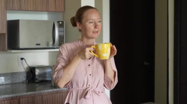 Genç bir kadın mutfakta bir fincan çay hazırlıyor ve onu içmekten zevk alıyor. Sıcak ve davetkar bir yuva ortamında rahatlatıcı bir öz bakım, rahatlama ve farkındalık anı.