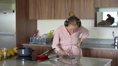 Genç bir kadın mutfakta yemek pişirirken neşeyle dans ediyor ve şarkı söylüyor. Sıcak bir ev ortamında yemek hazırlarken yaratıcılığın, kendini ifade etmenin ve keyif almanın neşeli ve neşeli bir anı.