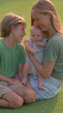 Genç bir anne, yemyeşil bir çimenlikte büyük oğlu ve yeni doğmuş çocuğuyla güneşli bir günün tadını çıkarır. Sevgi, bağ ve açık hava eğlencesiyle dolu neşeli bir aile anı ebeveynliği ve çocukluğu kutluyor.