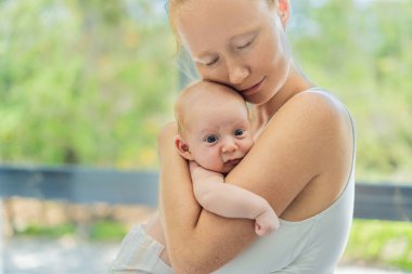 Anne evde bebeğine nazikçe sarılıyor, sıcak ve sevgi dolu bir anı paylaşıyor. Ebeveynlik, aile bağı ve anne sevgisi kavramı..