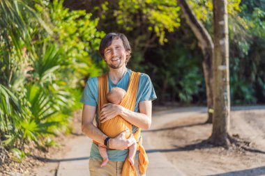 Babam küçük bebeğini parkta sarı bir askıda tutuyor. Sıcak ve sevgi dolu bir aile anı. Bebek giyimi, babalık ve ebeveyn-çocuk bağlama kavramı..