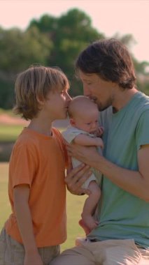Genç bir baba, yemyeşil bir çimenlikte büyük oğlu ve yeni doğmuş çocuğuyla güneşli bir günün tadını çıkarır. Sevgi, bağ ve açık hava eğlencesiyle dolu neşeli bir aile anı ebeveynliği ve çocukluğu kutluyor.