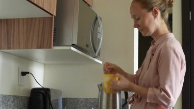 Genç bir kadın mutfakta bir fincan çay hazırlıyor ve onu içmekten zevk alıyor. Sıcak ve davetkar bir yuva ortamında rahatlatıcı bir öz bakım, rahatlama ve farkındalık anı.