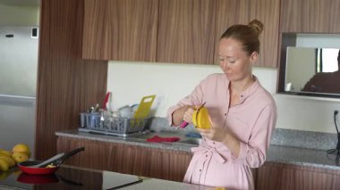 Genç bir kadın mutfakta duruyor, taze mangonun tadını çıkarıyor. Sağlıklı beslenmenin, tropikal lezzetlerin ve sıcak bir yuva ortamında ihtiyatlı yaşamanın basit ve canlı bir anı.