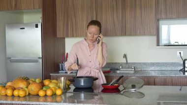 Mutfakta yemek pişirirken cep telefonuyla konuşan bir kadın. Meşgul ama rahat bir ev ortamında iletişim ve yemek hazırlama için uygun bir an.