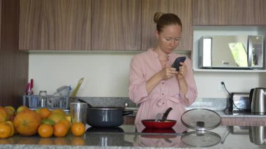 Genç bir kadın mutfakta duruyor, bir şey okurken akıllı telefon ekranına odaklanıyor. Konforlu bir ev ortamında birden fazla iş, teknoloji kullanımı ve modern yaşam tarzı ile ilişkilendirilebilir bir an