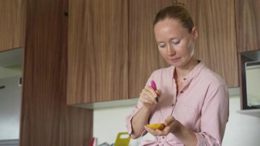 Genç bir kadın mutfakta duruyor, taze mangonun tadını çıkarıyor. Sağlıklı beslenmenin, tropikal lezzetlerin ve sıcak bir yuva ortamında ihtiyatlı yaşamanın basit ve canlı bir anı.