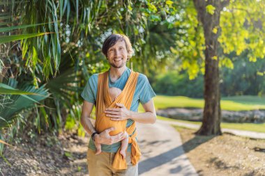 Babam küçük bebeğini parkta sarı bir askıda tutuyor. Sıcak ve sevgi dolu bir aile anı. Bebek giyimi, babalık ve ebeveyn-çocuk bağlama kavramı..